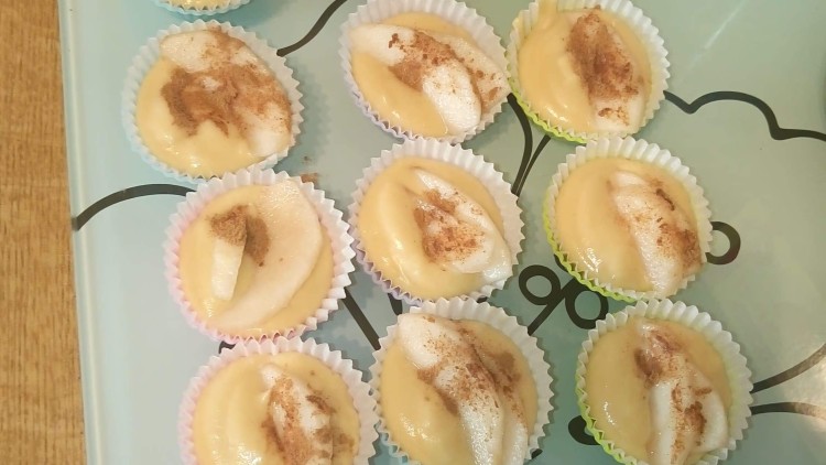 Mehrere ungebackene Muffins mit Teigfüllung, Birnenscheiben und Zimt in bunten Papierförmchen auf einer Glasplatte.