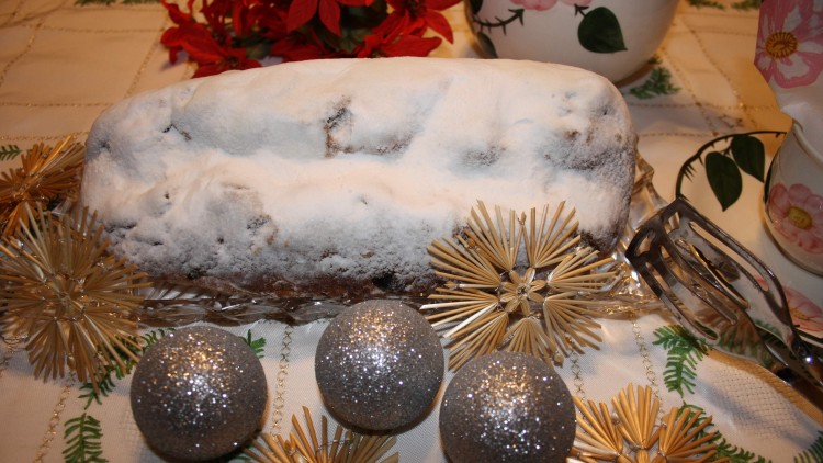 Ein mit Puderzucker bestäubter Christstollen liegt auf Glasplatte, dekoriert mit Glitzerkugeln, Strohsternen und rotem Weihnachtsstern.