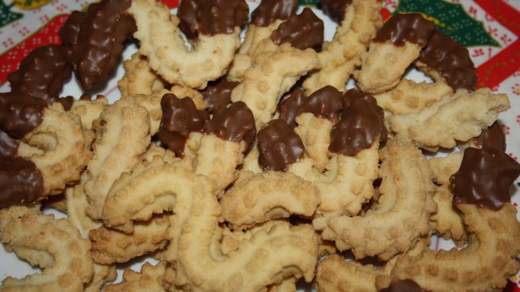 Plätzchenteller mit gebogenen Spritzgebäckstücken, teilweise mit Schokoladenüberzug, auf weihnachtlicher Tischdecke.