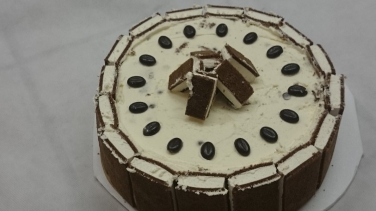Kaffeetorte mit heller Creme, Rechteckstücken an den Seiten, Schokoladendekor und Kaffeebohnen auf der Oberseite.