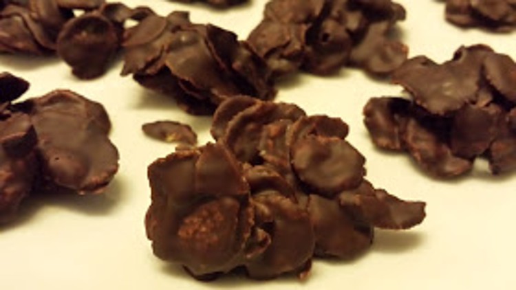 Schokoladenüberzogene Cornflakes-Häufchen liegen verteilt auf einer hellen Oberfläche.