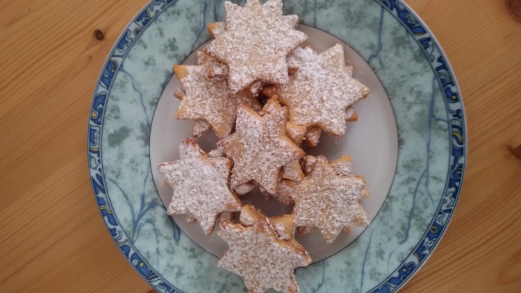 Plätzchen in Sternform mit Puderzucker liegen auf einem Teller mit grün-blauer Randverzierung auf einem Holztisch.