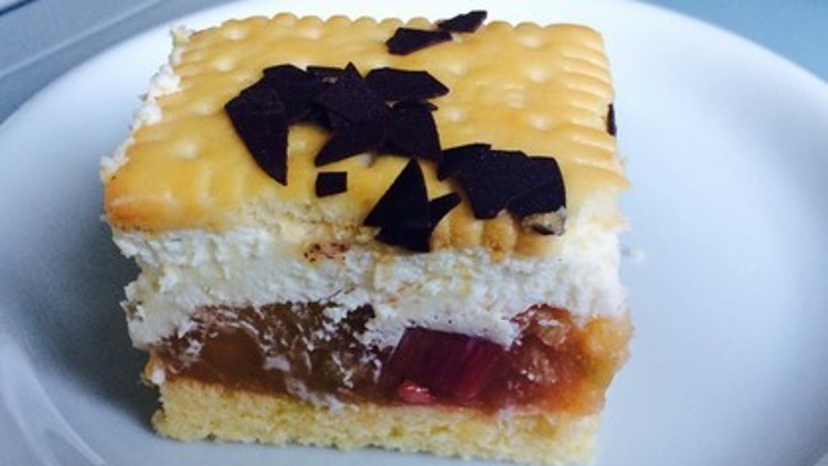 Ein Stück Kuchen mit Schichten aus Rhabarber, Sahne, Keks und Schokoraspeln liegt auf einem weißen Teller.