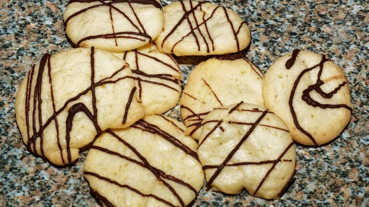 Mehrere runde, helle Kekse mit dünnen Linien aus dunkler Schokolade liegen auf einer gemusterten Steinoberfläche.