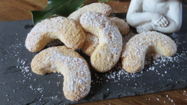 Vanillekipferl mit Puderzucker auf einer dunklen Schieferplatte, dahinter Stechpalmenzweig mit roten Beeren und Dekofigur.