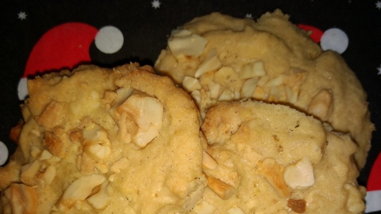 Drei Kekse mit grob gehackten Mandeln liegen auf einer schwarzen Serviette mit Weihnachtsmotiv aus roten Mützen und Sternen.