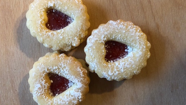 Drei runde Plätzchen mit Puderzucker und herzförmiger Öffnung, gefüllt mit roter Marmelade, auf hellem Holzuntergrund.