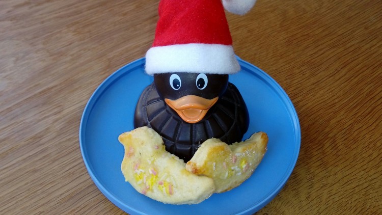 Eine schwarze Schokoente mit roter Weihnachtsmütze steht auf einem blauen Teller neben zwei Keksen mit Zuckerstreuseln.