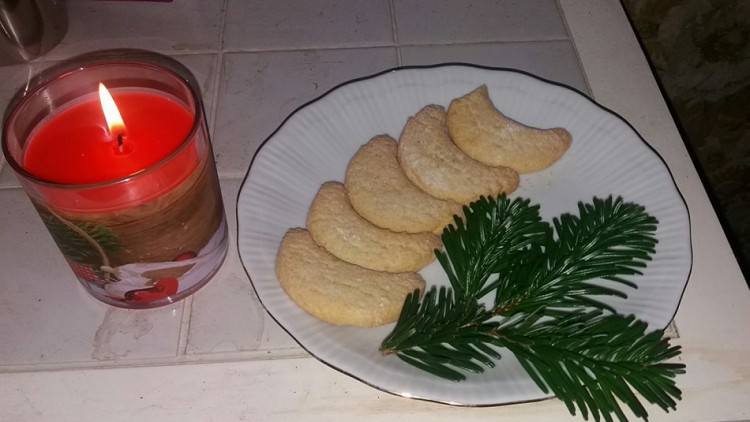 Ein weißer Teller mit Vanillekipferln und Tannenzweig, daneben eine rote Kerze in einem Glas mit Weihnachtsmotiv.