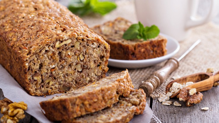 Ein Nussbrot mit sichtbaren Walnüssen liegt auf einem Holzbrett, daneben einzelne Brotscheiben, Walnüsse und Haferflocken.