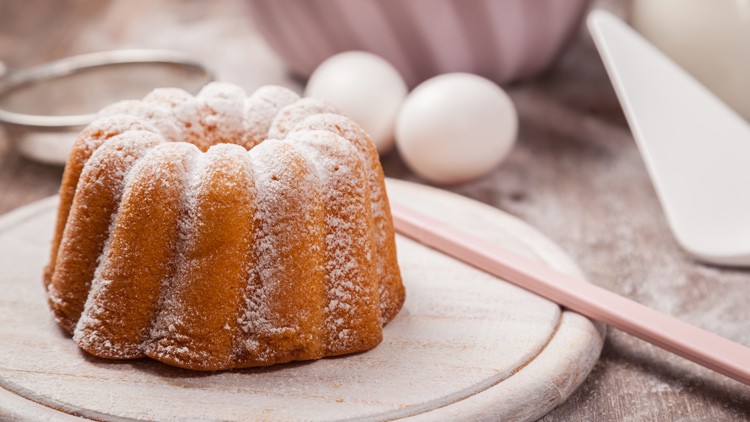 Ein kleiner Gugelhupfkuchen mit Puderzucker liegt auf einem runden Holzbrett, daneben Eier und ein rosa Backutensil.