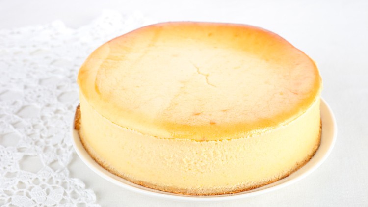 Runder Käsekuchen mit goldbrauner Oberfläche auf einem weißen Teller, daneben liegt ein weißes Häkeldeckchen.