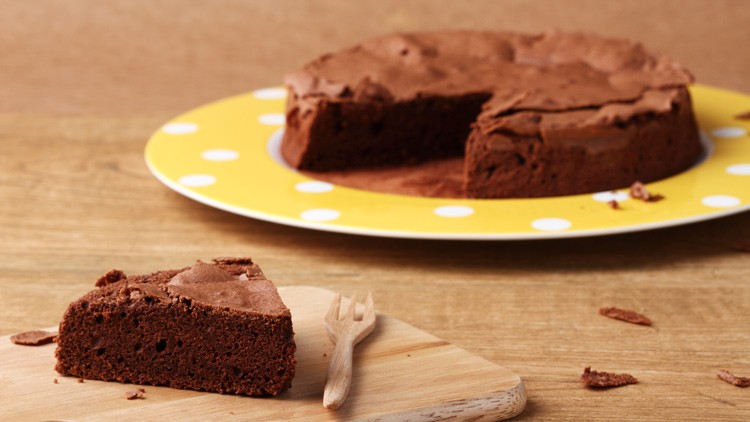 Ein Schokoladenkuchen auf gelbem Teller mit weißen Punkten, ein Stück davon liegt auf Holzbrett mit kleiner Holzgabel.