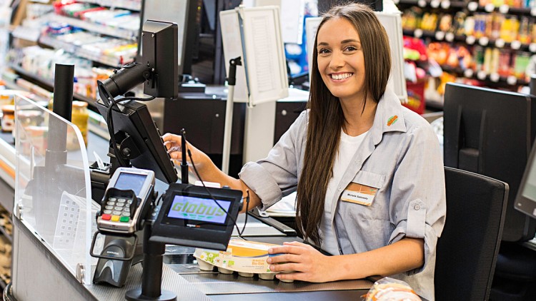 Person an einer GLOBUS-Kasse mit Scanner, Kartenterminal, Monitoren und Waren wie Eiern und Brot im Hintergrund.