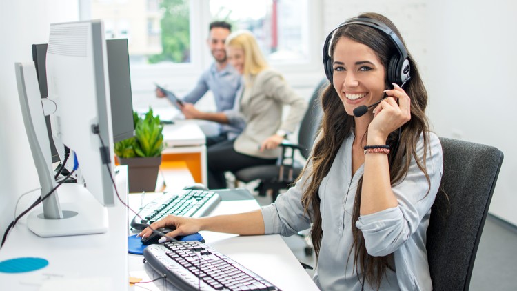 Person mit Headset und grauem Hemd sitzt an einem Schreibtisch mit Tastatur, Maus, Monitor und einer Pflanze.