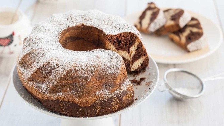 Ein Marmorkuchen mit Puderzucker auf einer weißen Tortenplatte, daneben ein Sieb und ein Teller mit Kuchenstücken.