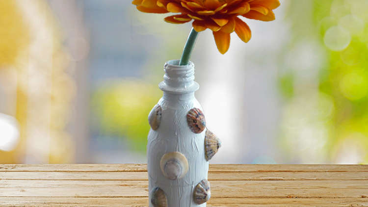 Eine weiße Glasflasche mit Muscheldekoration steht auf einem Holztisch und enthält eine orangefarbene Blume mit grünem Stiel.