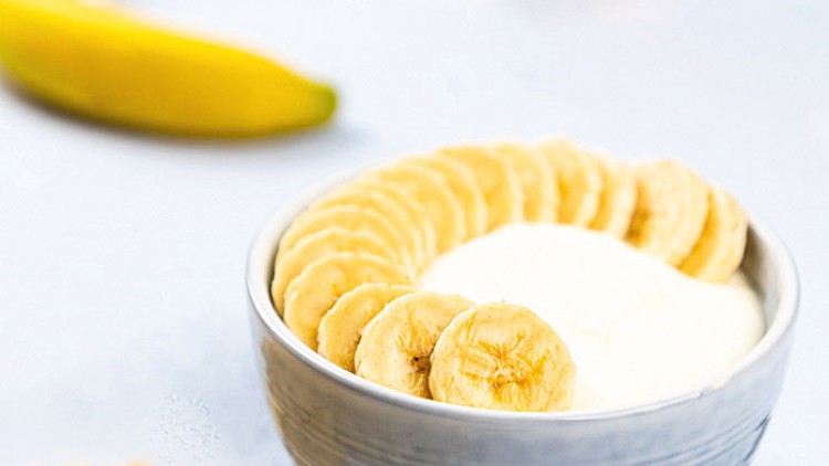 Graue Schale mit Joghurt und Bananenscheiben, daneben eine geschälte Banane und ein Löffel auf hellem Untergrund.