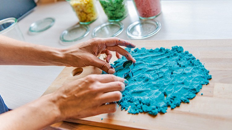 Hände formen blaue Knetmasse auf einem Holzbrett, im Hintergrund stehen Gläser mit gelber, grüner und roter Knetmasse.