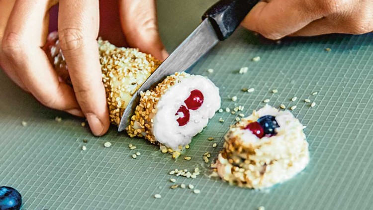 Hände schneiden mit einem Messer Fruchtsushi aus Sesamkruste und Reis auf grünem Schneidebrett, daneben Beeren.