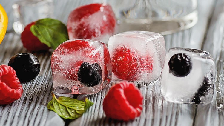 Eiswürfel mit eingefrorenen Himbeeren, Blaubeeren und Minzblättern liegen auf einer Holzfläche neben frischen Beeren.