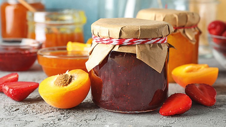 Verschlossene Marmeladengläser mit braunem Papierdeckel, frische Erdbeeren und Aprikosenhälften auf einem grauen Untergrund.