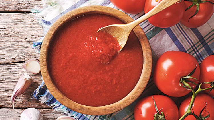 Rote Tomatensauce in einer Holzschale mit Holzlöffel, daneben frische Tomaten und Knoblauch auf einem rustikalen Untergrund.