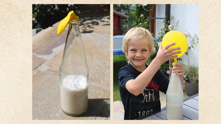 Eine Glasflasche mit weißer Flüssigkeit und einem gelben Ballon darauf, daneben Hände, die den gelben Ballon halten.