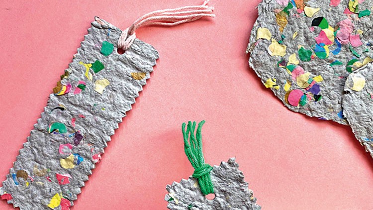 Graue, handgeschöpfte Papierstücke mit bunten Faser-Einschlüssen, davon zwei als Lesezeichen mit Kordel verziert, auf rosa Untergrund.
