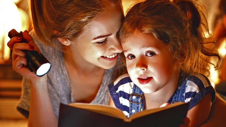 Person hält eine Taschenlampe, die auf ein geöffnetes Buch leuchtet, im Hintergrund ein Kuscheltier und warme Beleuchtung.