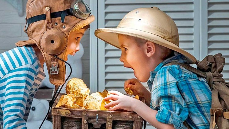 Zwei Kinder in Abenteurerkostümen betrachten eine Schatzkiste mit goldfarbenen Steinen, im Hintergrund ein Raum mit Holzdetails.