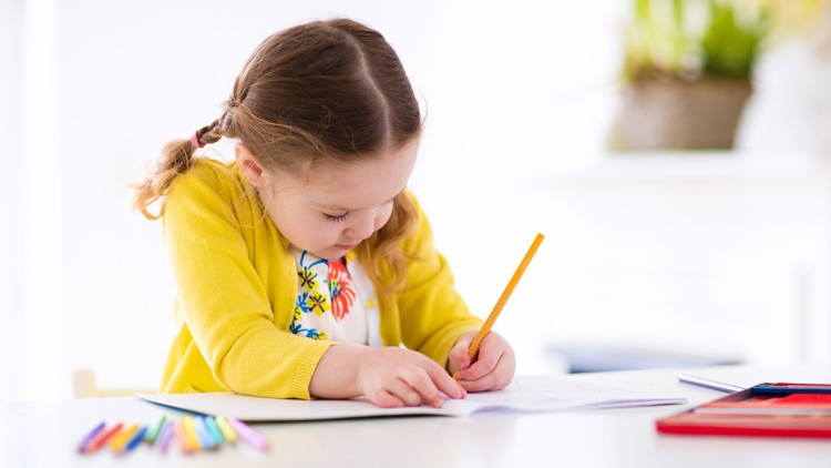 Ein Kind in gelbem Pullover malt mit einem gelben Bleistift auf Papier, daneben liegen bunte Stifte auf einem weißen Tisch.