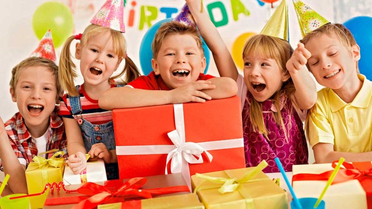 Kinder mit Partyhüten sitzen an einem Tisch mit bunten Geschenken, Luftballons und einer \