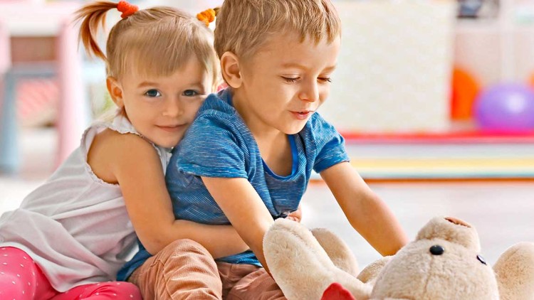 Zwei Kinder sitzen auf einem Teppich in einem farbenfrohen Raum mit Spielzeug, im Vordergrund ein Plüsch-Teddybär.