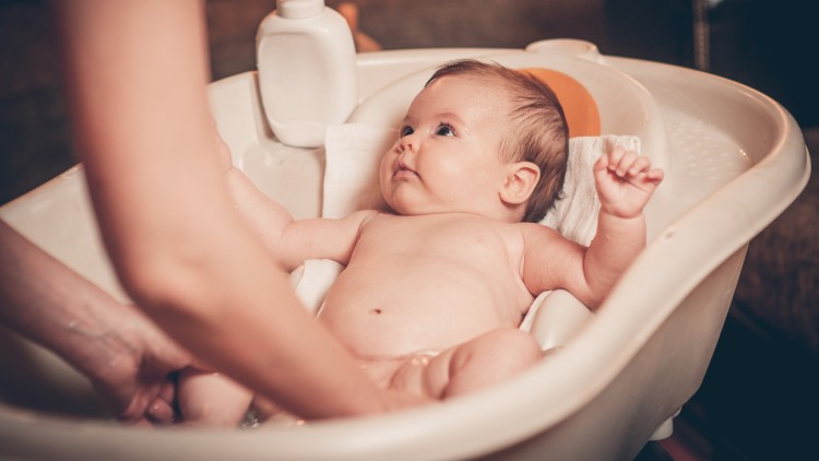 Ein Baby liegt in einer weißen Babywanne mit einem Handtuch im Hintergrund und einer Lotionflasche am Wannenrand.