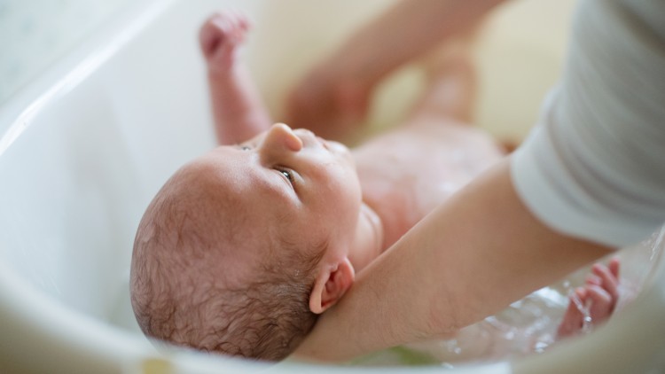 Ein Baby mit nassen Haaren wird von einer Person mit einer Hand gestützt und in einer weißen Badewanne gebadet.