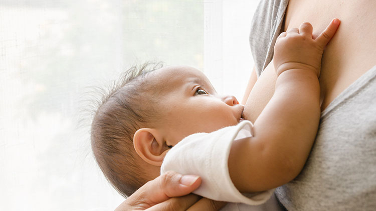 Ein Baby wird an der Brust einer Person gehalten, die einen grauen Stoff trägt, Hintergrund mit heller Gardine.