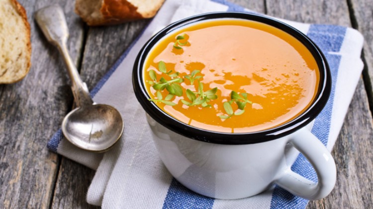 Eine weiße Tasse mit orangefarbener Suppe, garniert mit frischen Kräutern, steht auf einem Stofftuch neben einem Löffel und Brot.