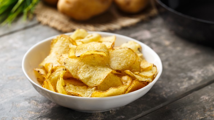 Eine weiße Schüssel mit goldgelben Kartoffelchips steht auf einem Holztisch, im Hintergrund liegen frische Kartoffeln.