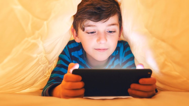 Kind in gestreiftem Pullover liegt unter einer Decke und hält ein leuchtendes Tablet mit beiden Händen.