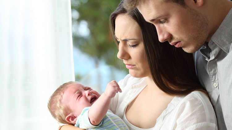 Person hält ein Baby in gestreiftem Strampler in den Armen, im Hintergrund sind helle Vorhänge und unscharfe Bäume sichtbar.