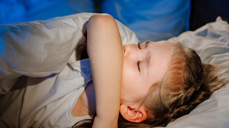 Person liegt in einem Bett mit weißer Bettwäsche, umgeben von blauem Licht, und hebt einen Arm über den Kopf.
