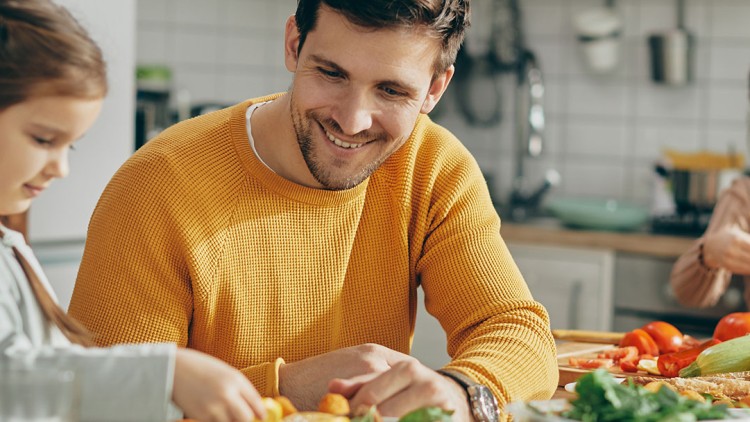 Personen sitzen an einem gedeckten Tisch mit Gemüse, Brot und Salat in einer Küche, eine Person faltet gelbes Tuch.