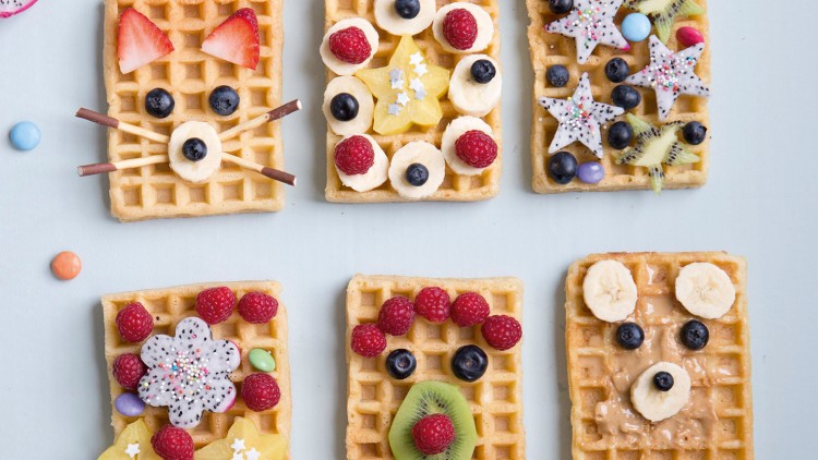 Sechs Waffeln mit buntem Obst wie Erdbeeren, Bananen, Kiwi, Blaubeeren und Himbeeren kreativ dekoriert auf hellem Untergrund.