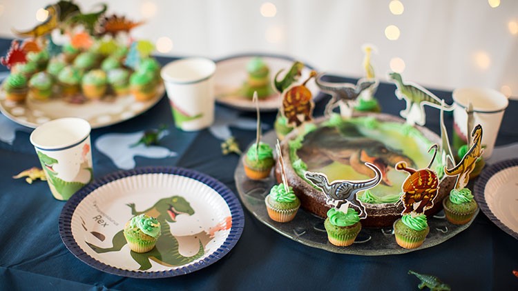 Eine festlich gedeckte Tafel mit Dinosaurier-Motiven, grünen Muffins und einem Dinosaurier-Kuchen auf dunkler Tischdecke.