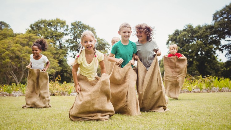 Mehrere Kinder hüpfen mit großen Jutesäcken auf einer grünen Wiese, im Hintergrund sind Bäume und Blumen zu sehen.