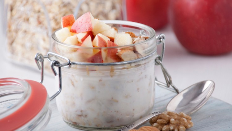 Ein Glas mit Müsli, Joghurt und Apfelstücken steht auf einem Holzbrett, daneben eine silberne Löffel und Nüsse.