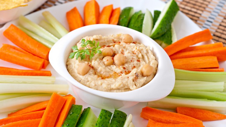 Ein Teller mit Hummus in einer Schale, dekoriert mit Kichererbsen und Petersilie, umgeben von Gurken-, Karotten- und Selleriesticks.
