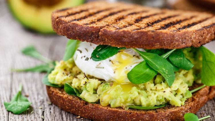 Geröstetes Vollkorn-Sandwich mit Avocadocreme, pochiertem Ei, Spinatblättern und Kräutern auf einem Holztisch.