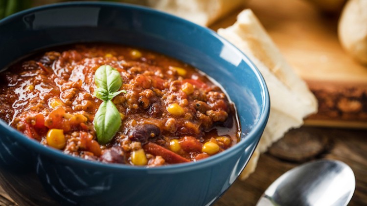 Eine blaue Schüssel mit Chili con Carne, garniert mit frischem Basilikum, daneben ein Löffel und frisches Brot auf einem Holzbrett.